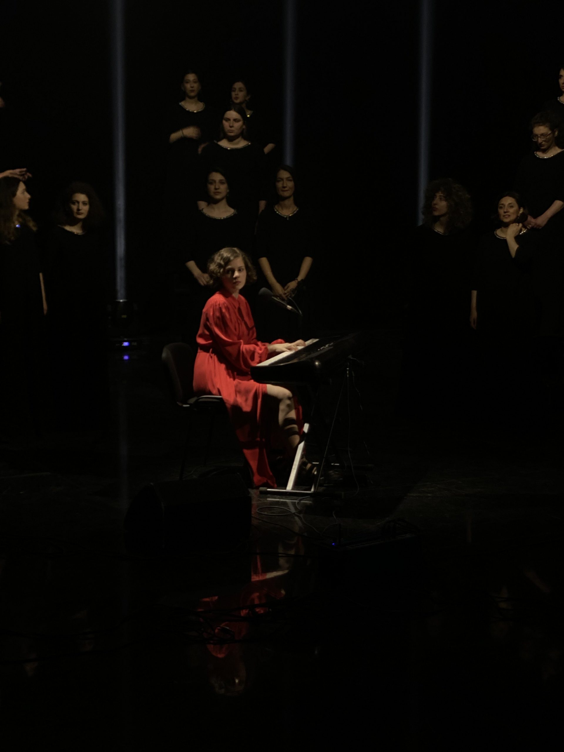 Lizi Gozalishvili spielt Klavier in rotem Kleid während einer Chor-Performance in Georgien, umgeben von Sängerinnen in schwarzen Kleidern